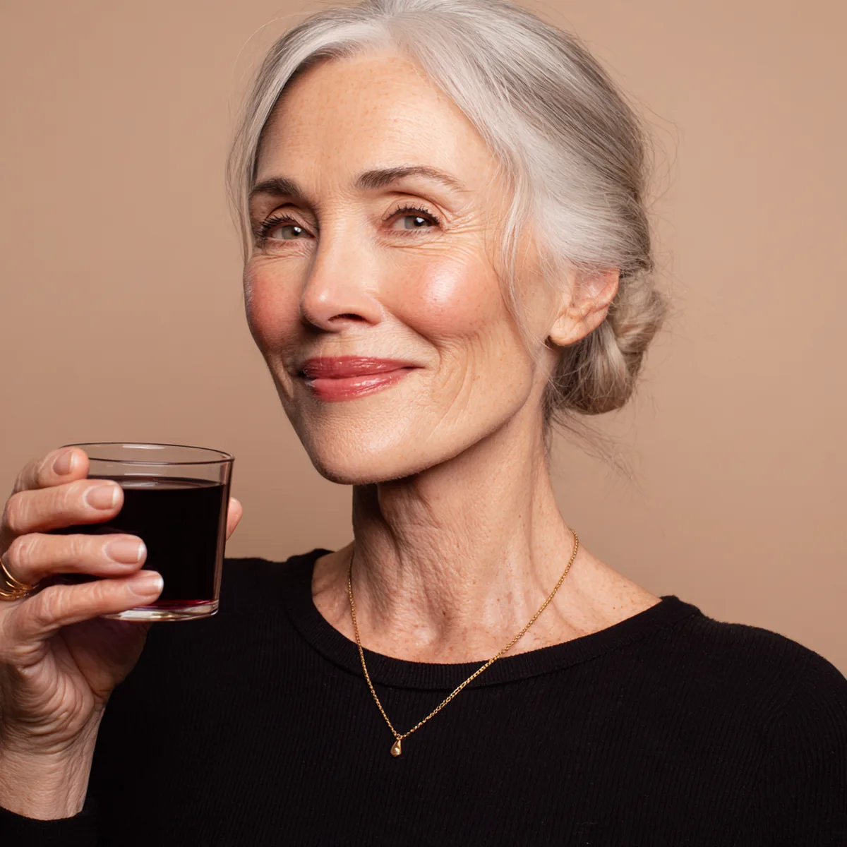Elegant silver-haired woman in a black top raising a small glass of collagen drink and smiling