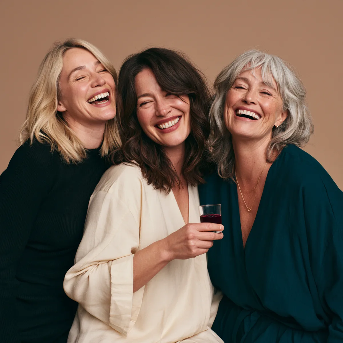 Three women in their forties and fifties laughing together, one holding a small glass of collagen drink