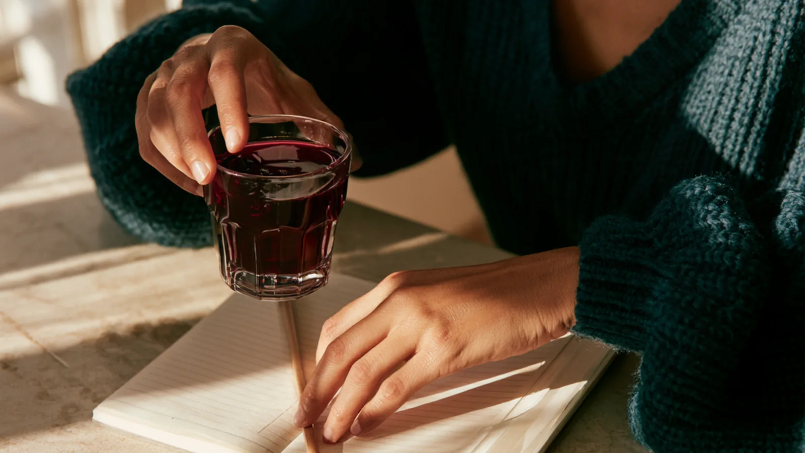 A woman's hands with a small collagen drink and a pencil on an open diary at a kitchen counter