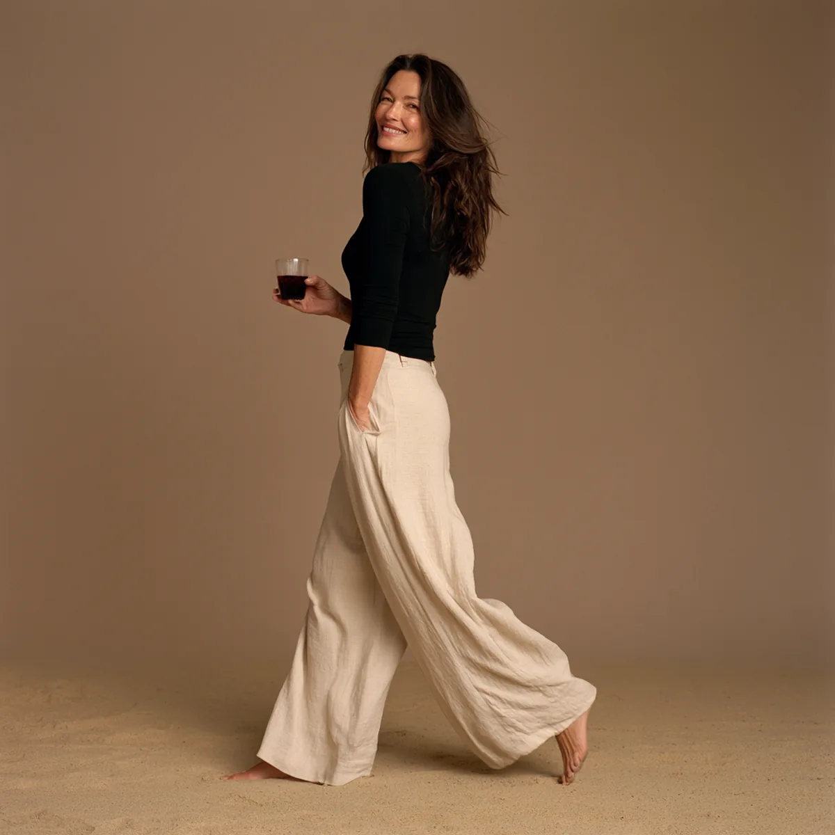 Woman in a black top and cream trousers walking barefoot, laughing back over her shoulder