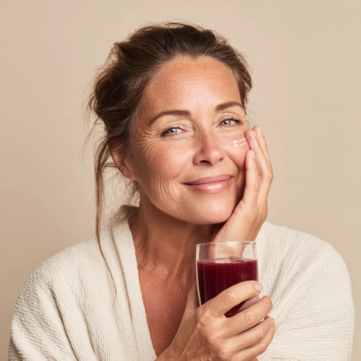 Woman dabbing face cream on her cheek while holding a small glass of collagen drink, smiling