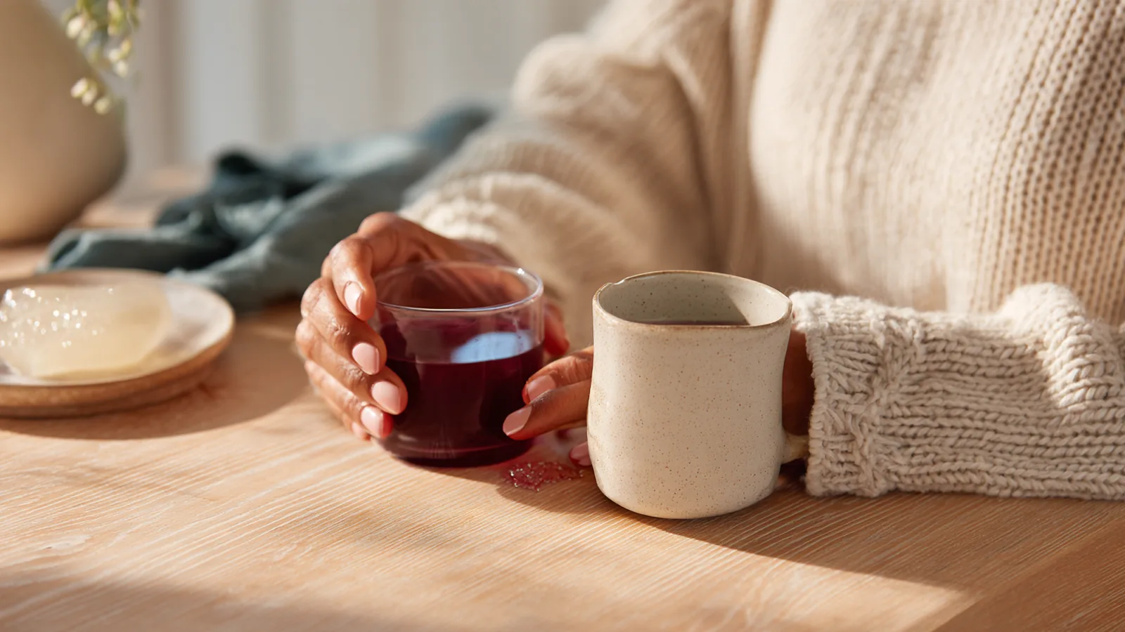 A woman's hands tilting a glass of berry-red collagen drink beside a steaming mug and a dish of gelatin jelly