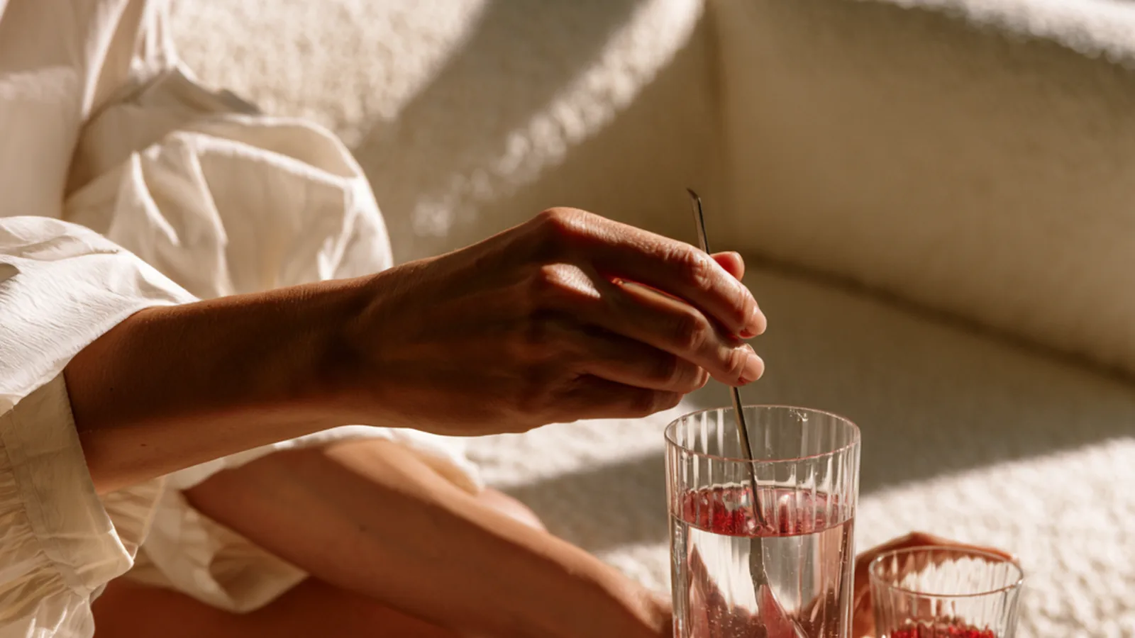 A woman's hands stirring liquid collagen into a tall glass of water, the pack beside it on an oak table