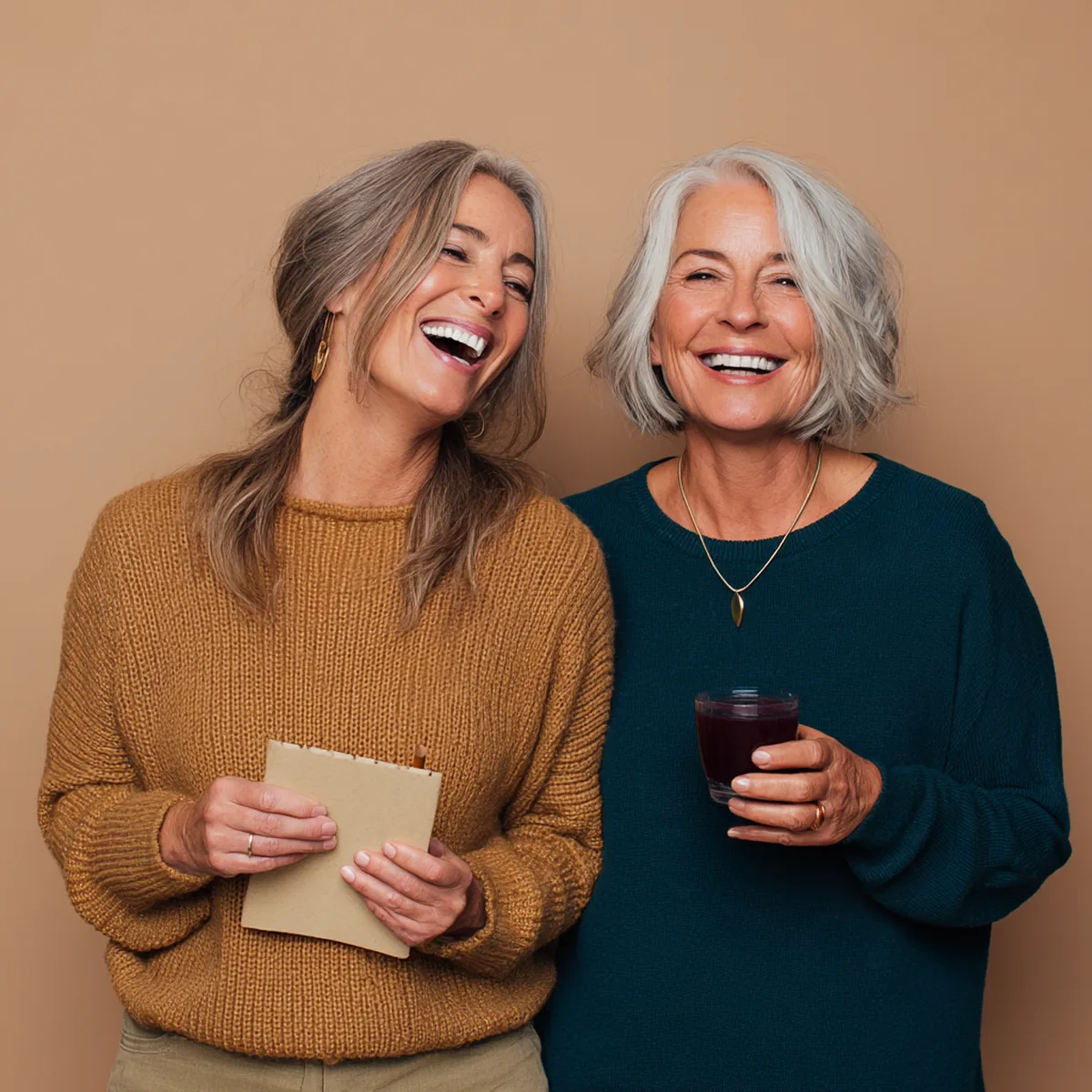 Two women laughing together, one holding a notepad and pencil, the other a small glass of berry-red drink