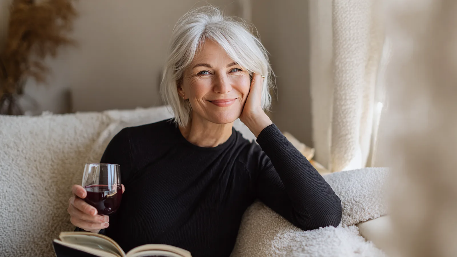 Woman in her forties on a cream sofa with a notebook, lifting a small glass of berry-red drink and smiling