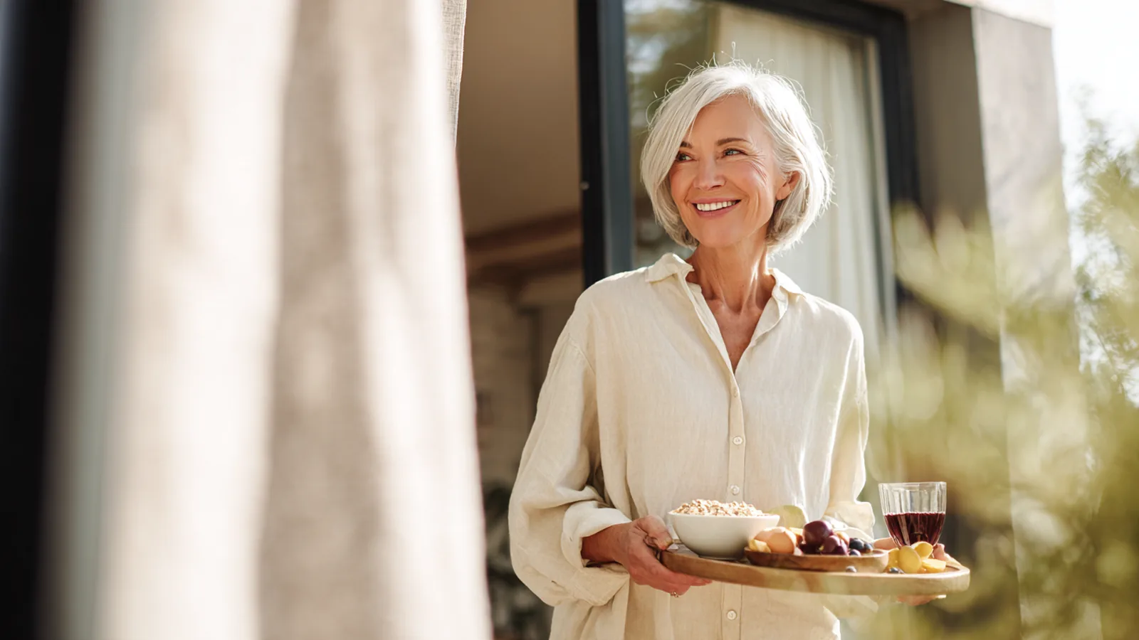 Silver-haired woman carrying a breakfast tray with a small berry-red drink out onto a terrace