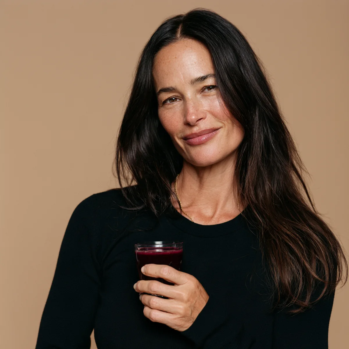 Dark-haired woman in her forties in a black top turning with a calm smile, holding a small berry-red drink