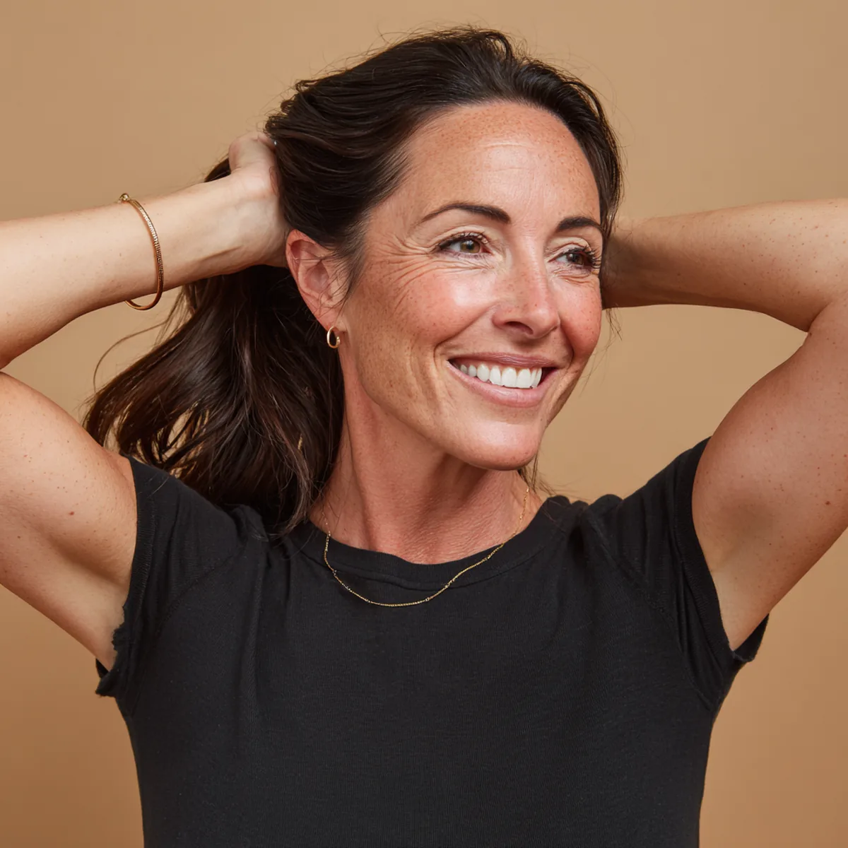 Woman in her late forties in a black tee laughing as she gathers her long dark hair into a ponytail