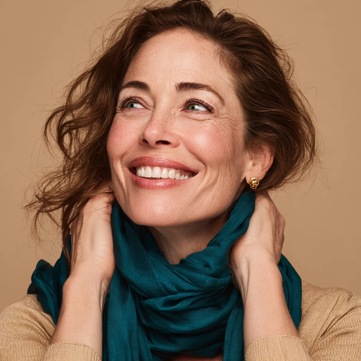 Laughing woman in her fifties with wavy brunette hair tying a deep teal silk scarf loosely at her neck