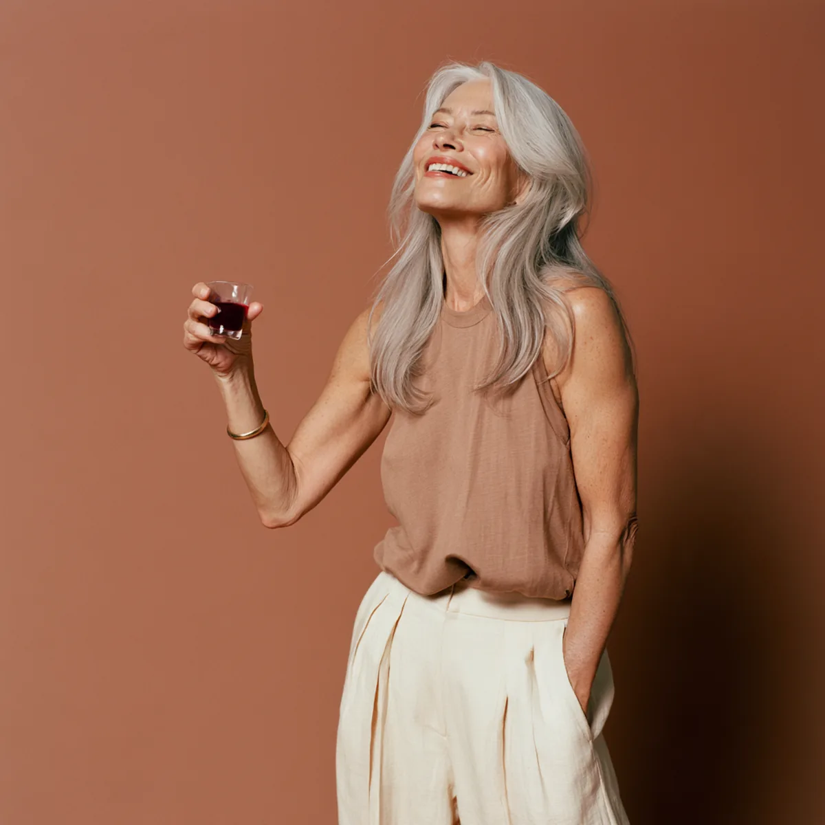 Silver-haired woman in her fifties standing tall and lifting a small glass of liquid collagen in a toast