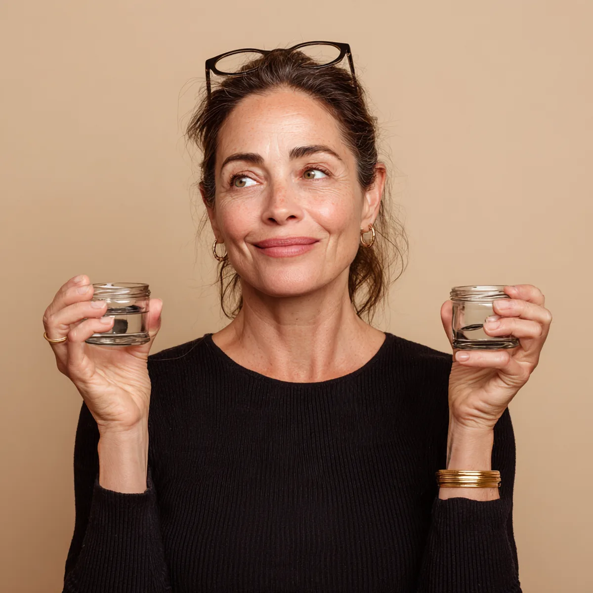 Woman in her forties holding a small plain glass jar in each hand and comparing them with a playful smile