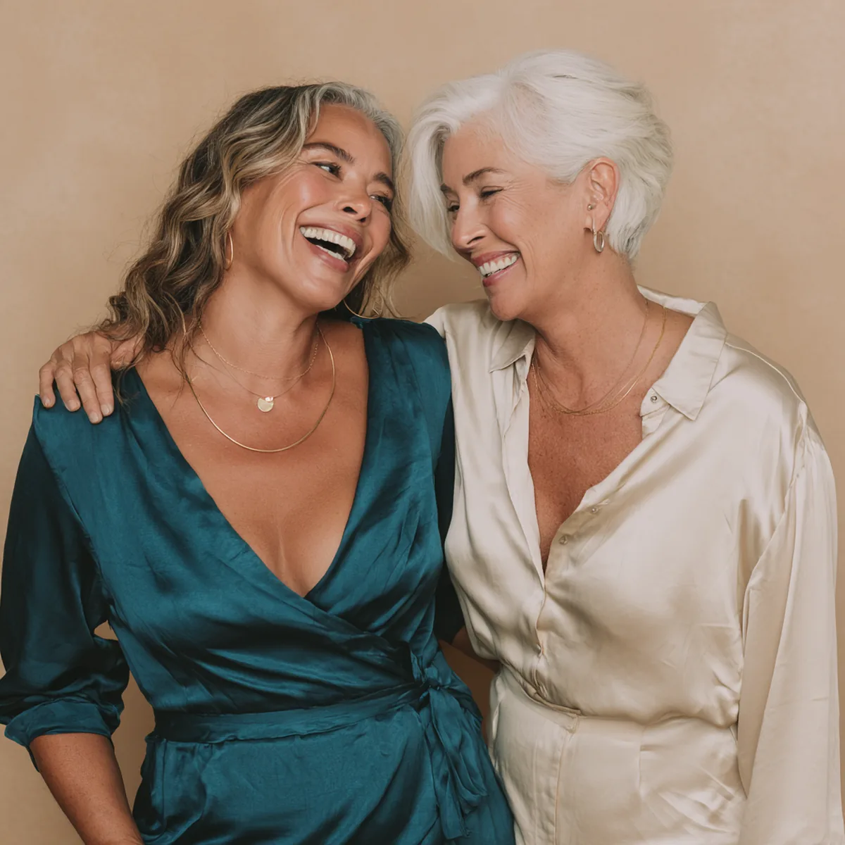 Two friends dressed for a celebration laughing as one fastens the other's gold necklace