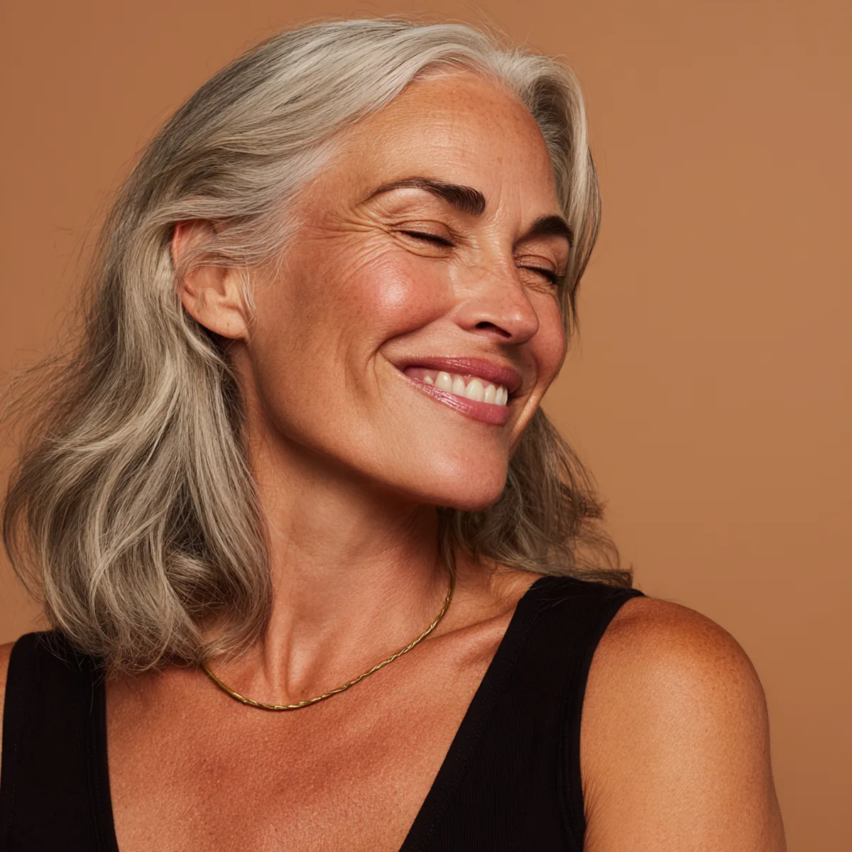 Silver-haired woman in her fifties laughing softly, luminous skin with fine natural smile lines