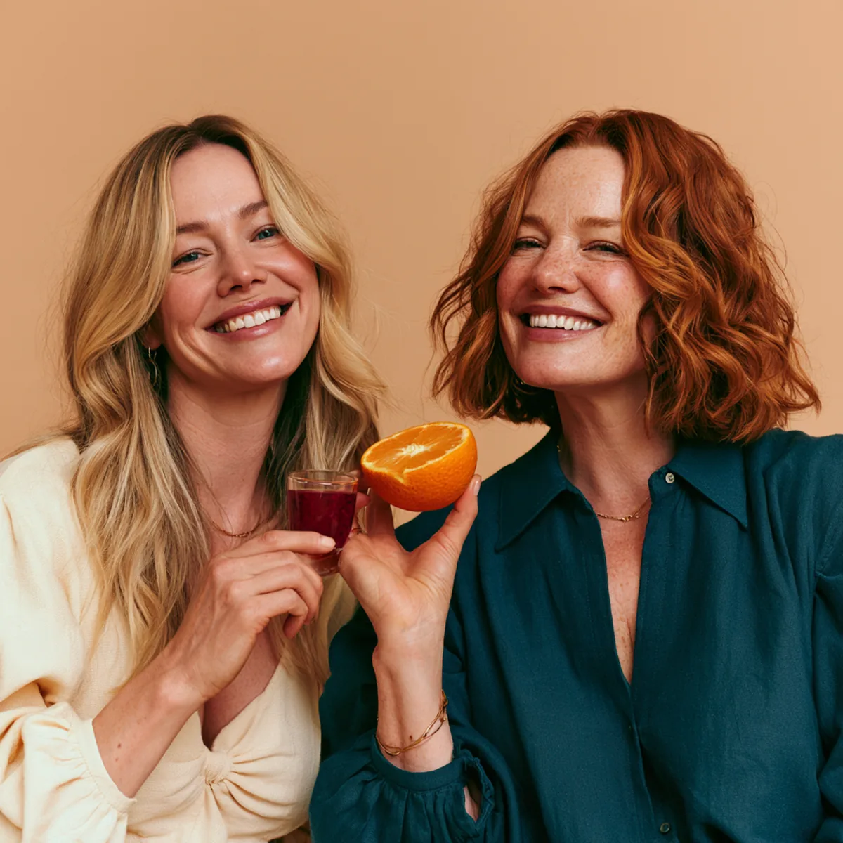 Two laughing women toasting with a small glass of collagen drink and half an orange